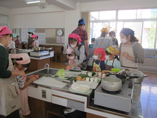 親子料理教室の写真です。子どもが野菜を切るのを後ろから母親が手助けしています